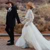A bride and groom in western attire holding hands in a desert landscape, with the bride in a flowing gown and the groom in a black suit and cowboy hat.