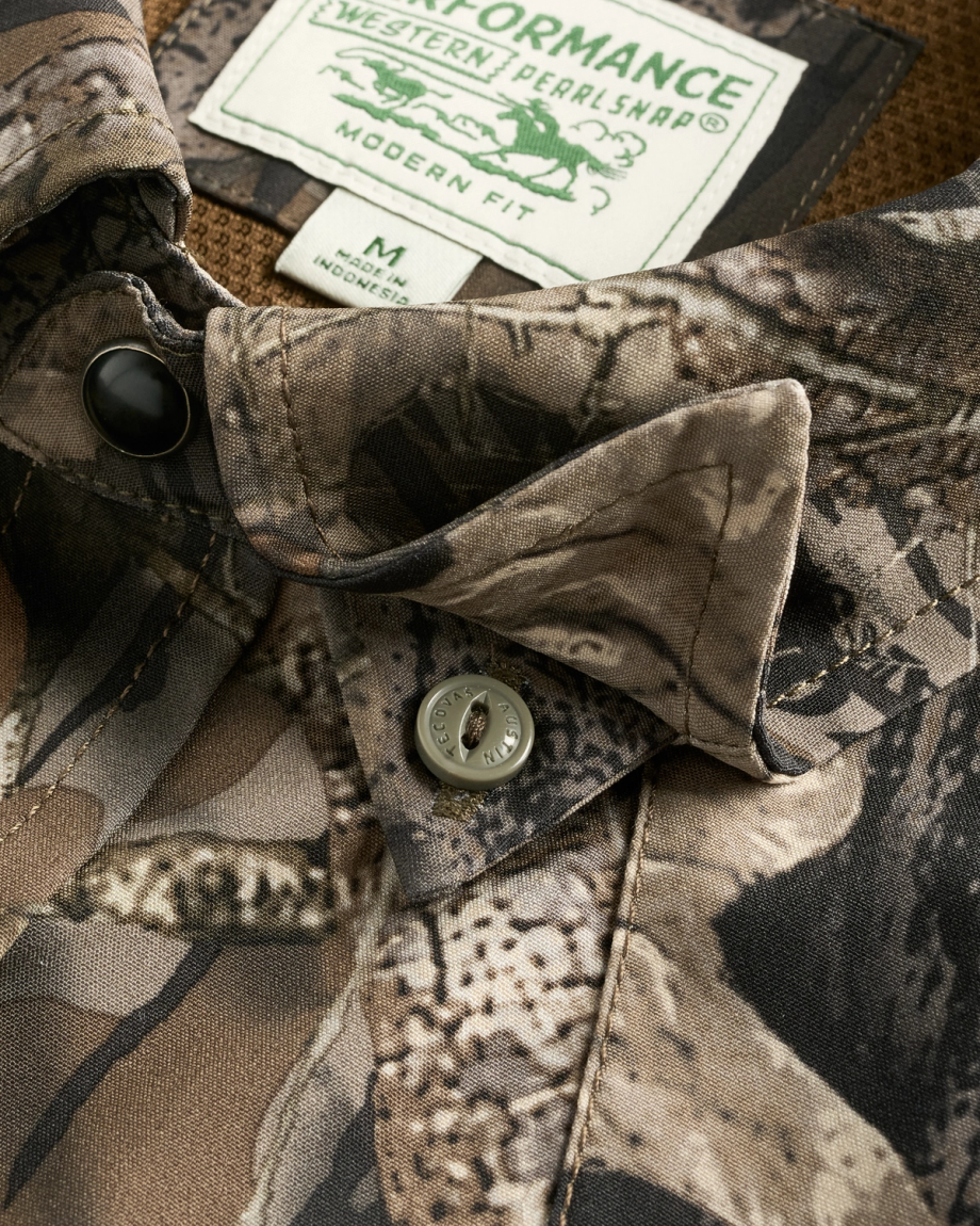 Close-up of a camouflage-patterned shirt with a buttoned collar and visible label reading “PERFORMANCE Western Pearl Snap, Modern Fit, M, Made in Bangladesh.”.