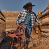 Man in denim and cowboy hat carries a saddle by a wooden fence and trailer, with dry hills in the background.