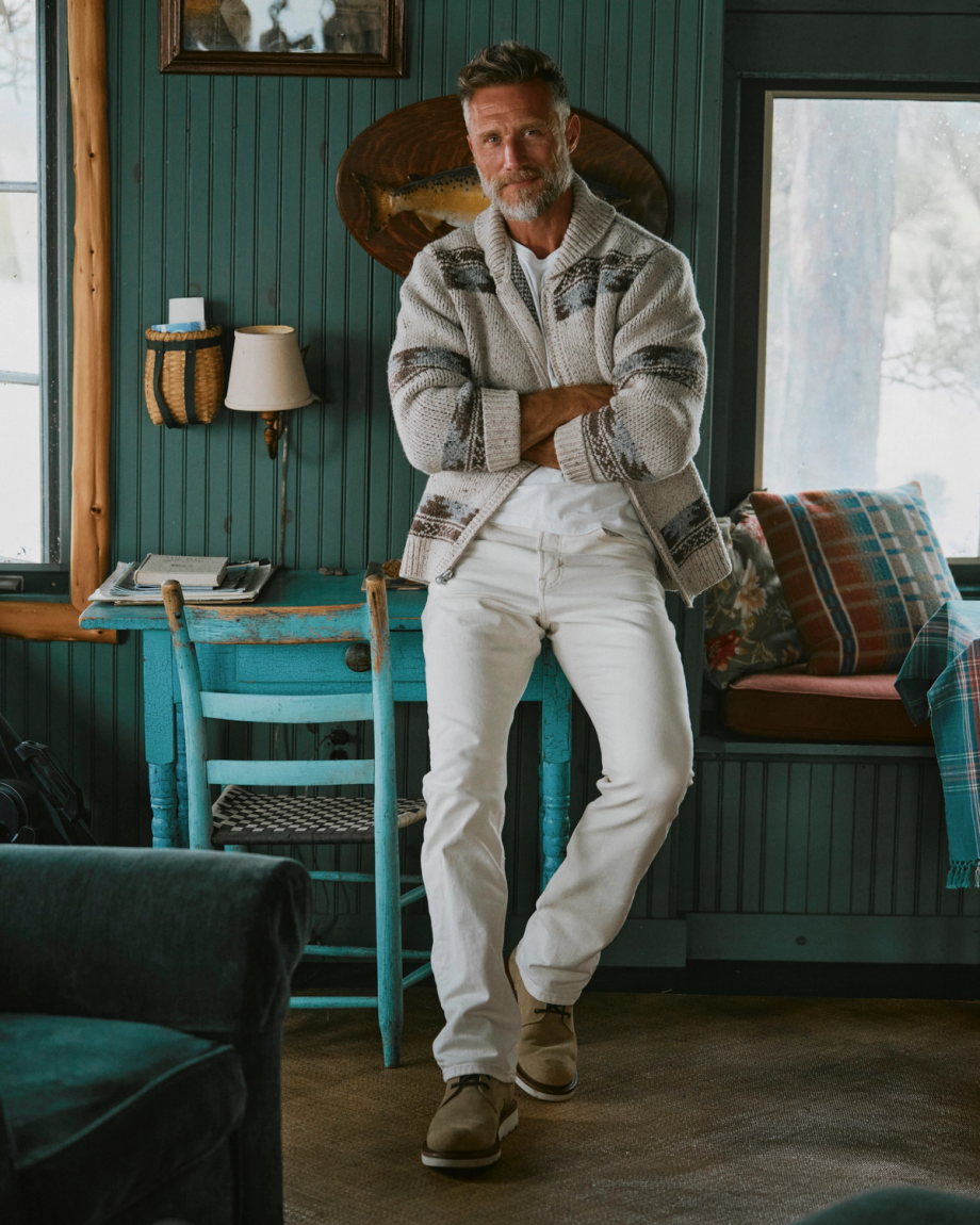 The Knox Lace Up Shearling in Sandstone Roughout. Man with gray hair and beard, wearing a patterned cardigan and white pants, stands casually indoors by a turquoise chair, arms crossed, with windows and sandstone roughout wooden furniture in the background.