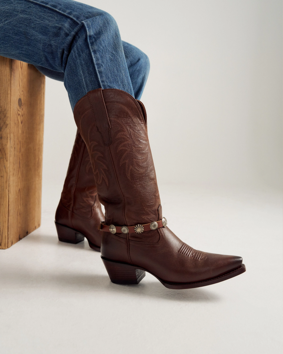 Person wearing blue jeans and brown leather cowboy boots with decorative stitching, seated on a wooden bench against a plain background.