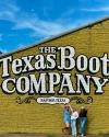 Three people pose in front of a large yellow brick wall painted with "The Texas Boot Company, Bastrop, Texas.
