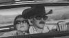A woman and a man wearing sunglasses sit in the front seat of a car; the man is driving and wearing a cowboy hat. The image is black and white.
