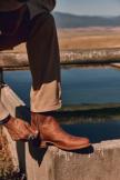Person zipping up a brown leather boot while sitting on a concrete ledge near a body of water.