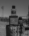 Black and white photo of Ranger Station fragrance products, including two bottles and a box, placed on a wooden fence post in an outdoor rural setting with a windmill visible in the background.