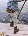 A person in jeans, a denim jacket, and boots stands on a dock holding a net over water with lily pads.
