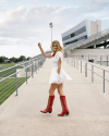 A person in a white dress and red boots stands on a walkway near a modern stadium with tiered seating and a cloudy sky in the background.