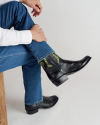 A person wearing blue jeans and black leather boots sits on a wooden bench with one leg crossed over the other.