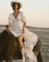 A woman in a white dress and hat rides a dark horse in a barren landscape, capturing a moment of serene motion.
