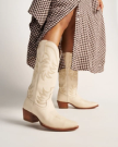 The Annie in Bone Cowhide. A person wearing bone cowhide cowboy boots and a brown checkered skirt stands against a plain background.