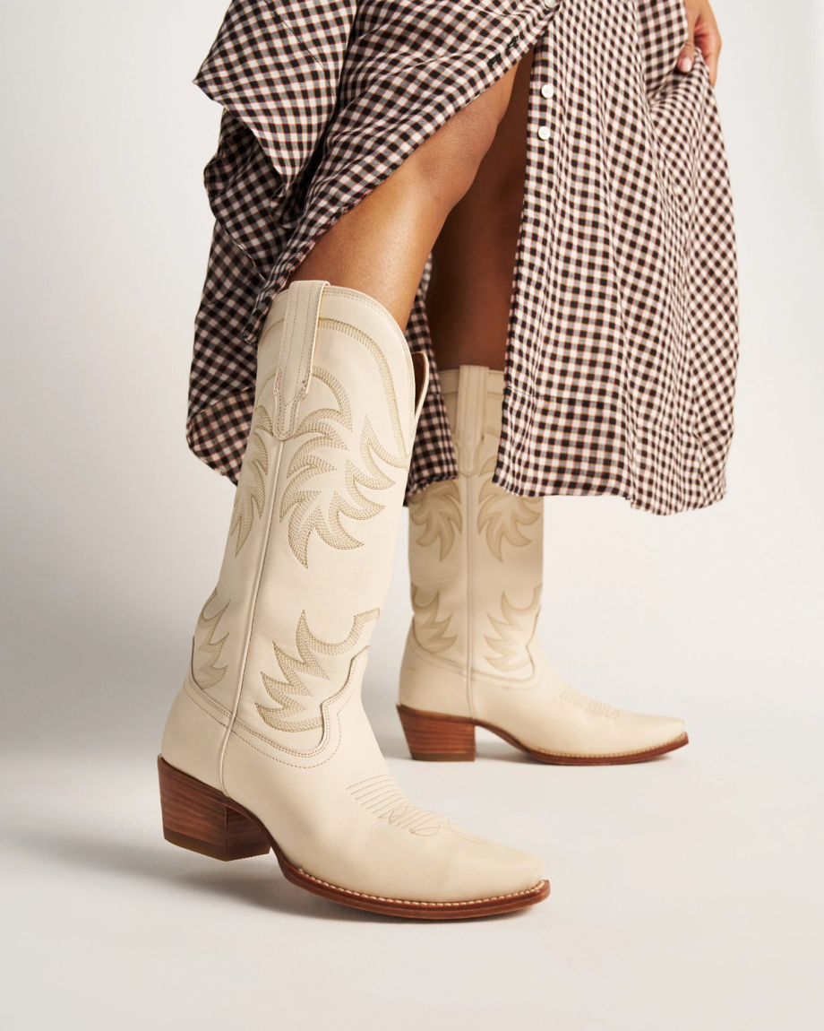 The Annie in Bone Cowhide. A person wearing bone cowhide cowboy boots and a brown checkered skirt stands against a plain background.