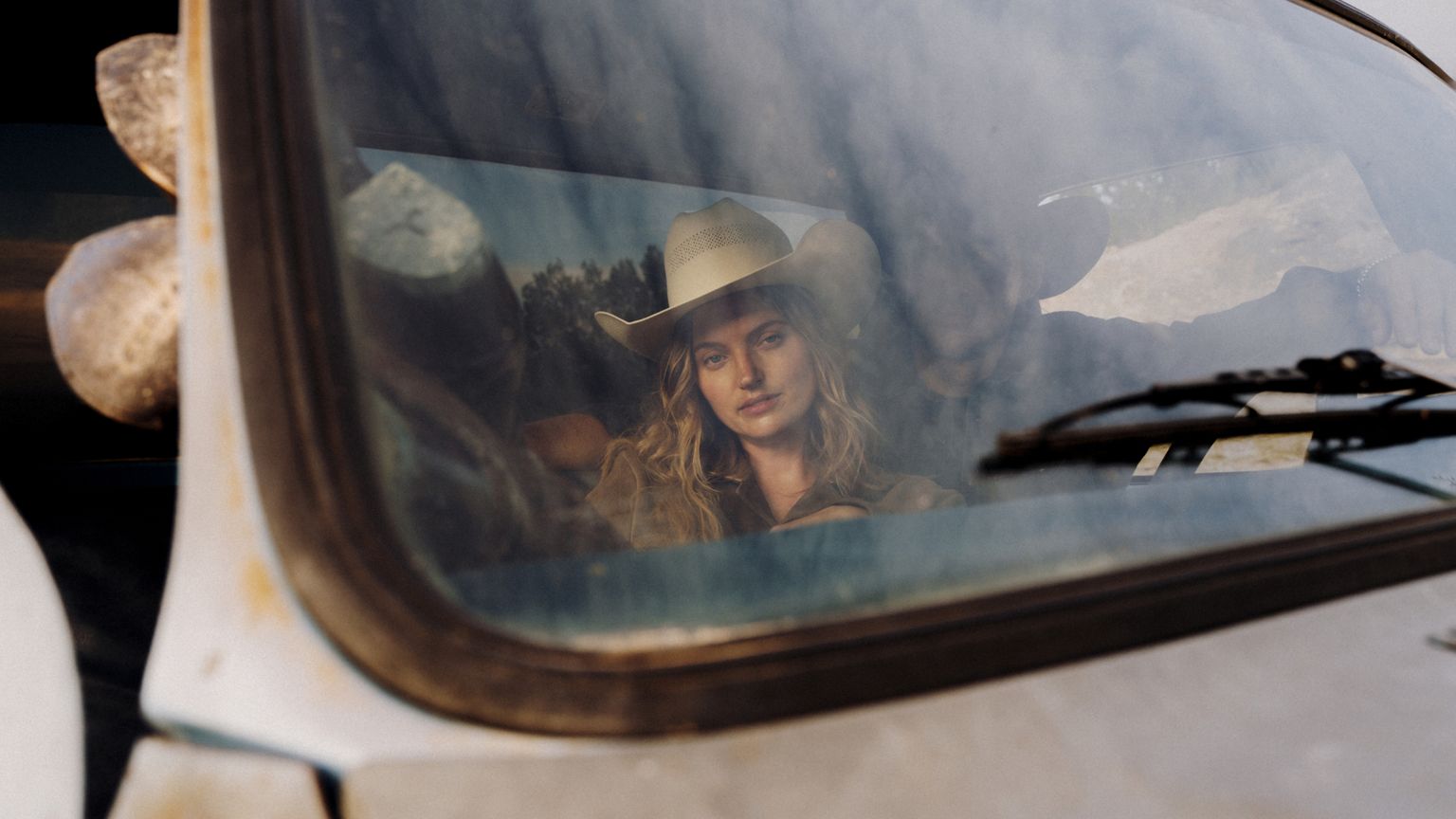 A woman wearing a cowboy hat sits in the passenger seat of a vehicle, looking out the front windshield. The image is taken from outside through the glass.