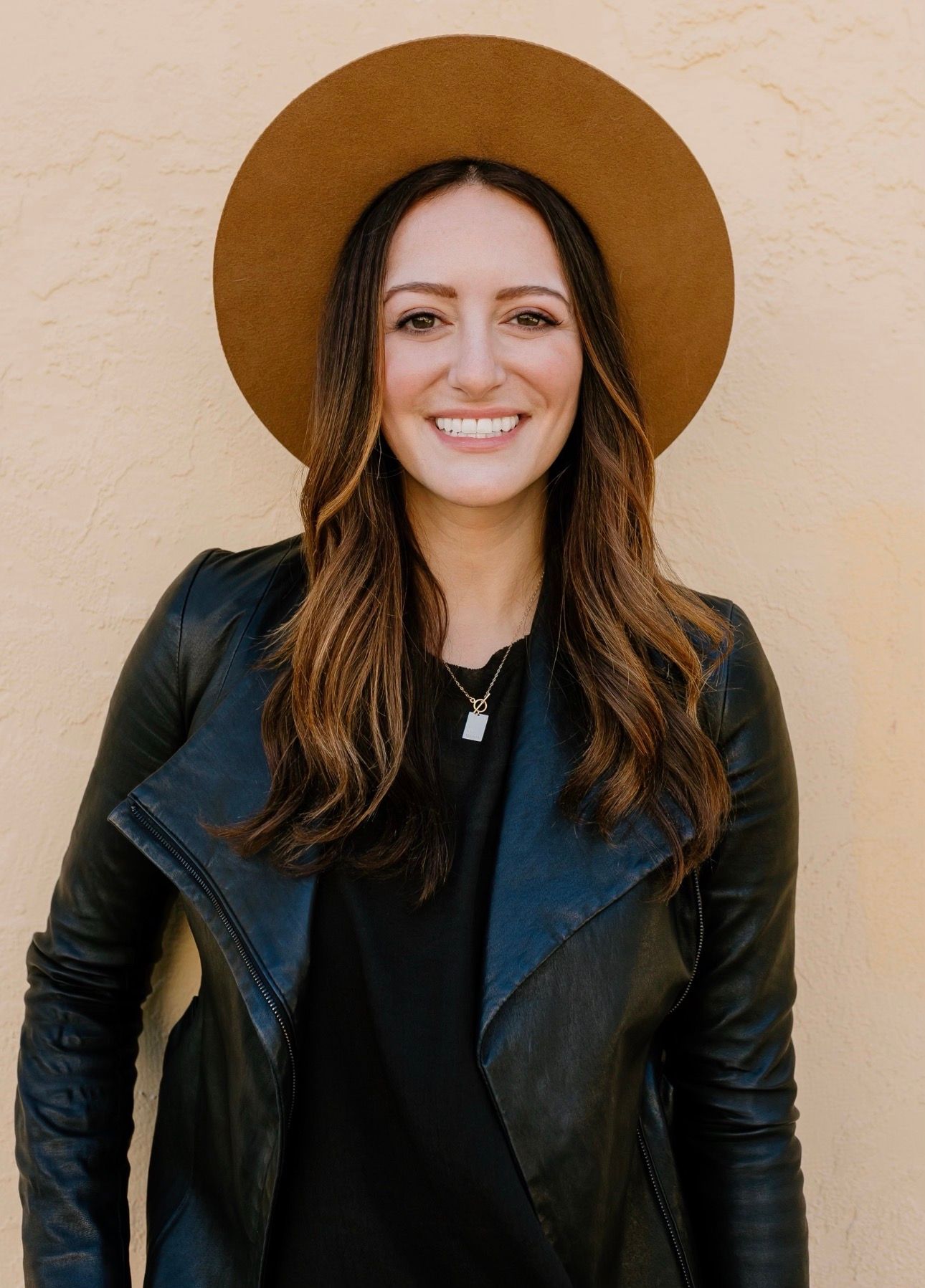 Woman with long brown hair wearing a tan hat, black leather jacket, and black shirt, standing against a light textured wall and smiling at the camera.