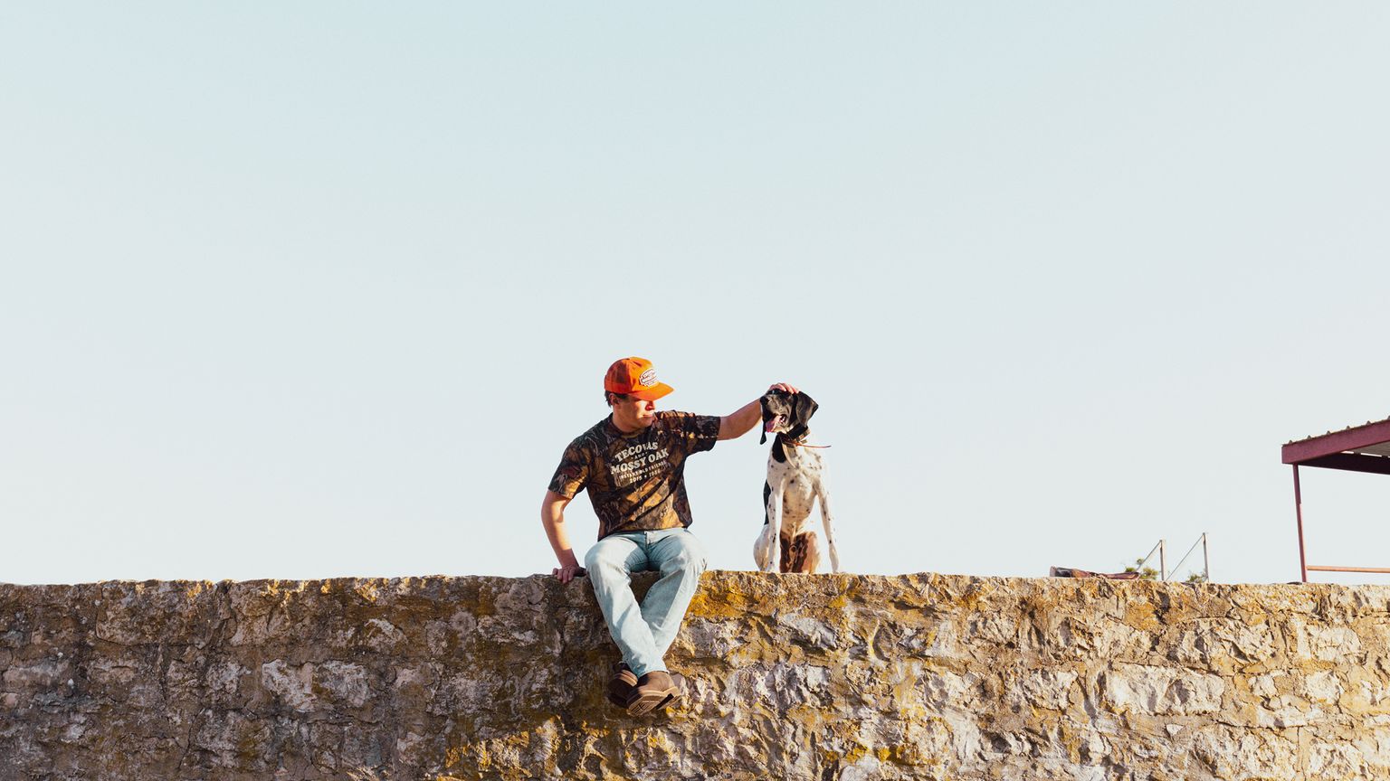 A person wearing an orange cap and camo shirt sits on a stone wall, petting a black and white dog beside them under a clear sky.