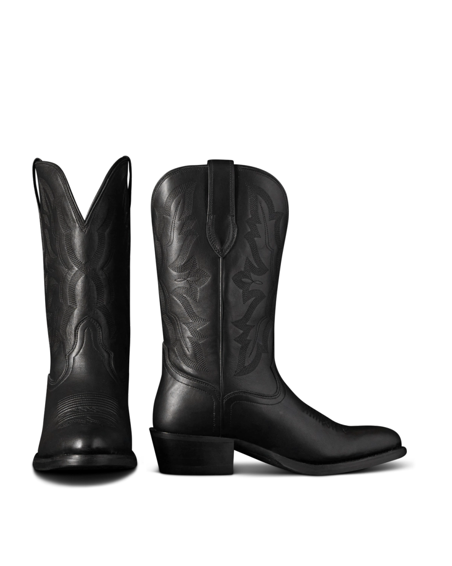 The Britt in Midnight Cowhide. Two black leather cowboy boots crafted from midnight cowhide with decorative stitching are shown; one boot faces forward, while the other is viewed from the side.
