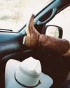 Cowboy boots rest on a car dashboard next to a white cowboy hat, with a desert landscape visible through the windshield.