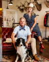 A man and woman pose indoors; the man sits on a red leather couch petting a black and white dog, while the woman stands behind him. Decor includes mounted deer heads and clothing racks.