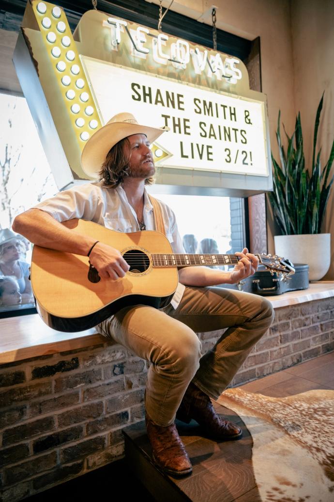 A musician in a white shirt and cowboy hat plays an acoustic guitar, sitting under a lit marquee sign announcing a live event.