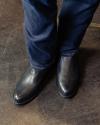 A close-up view of a person wearing dark blue jeans and shiny black leather boots. the feet are slightly angled on a dark floor.