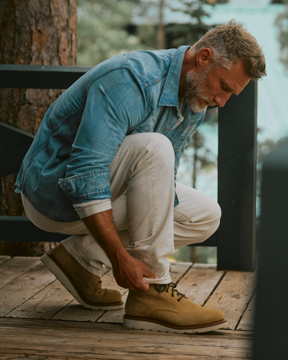 The Knox Lace Up Shearling in Sandstone Roughout. A man in a denim shirt and light pants bends down outdoors to tie the laces of his tan Knox Lace Up Shearling Roughout boots on a wooden deck.