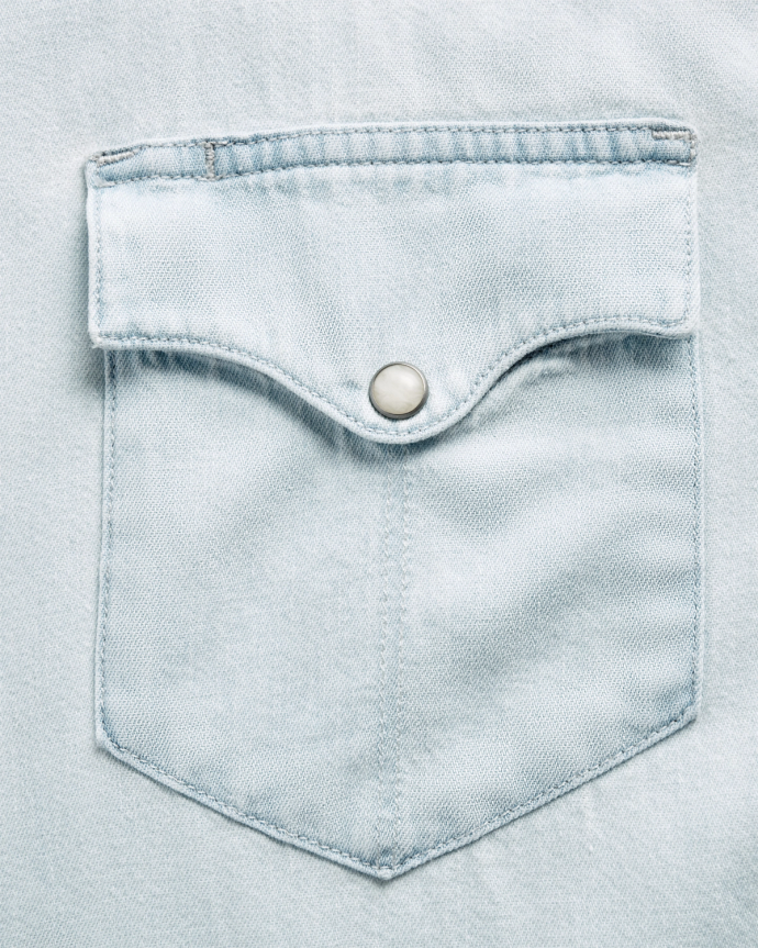 Close-up of a light blue denim shirt pocket with a flap and a metal snap button.