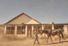 A cowboy leads a saddled horse past a weathered ranch house while other riders wait on the porch.
