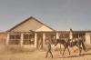A cowboy leads a saddled horse past a weathered ranch house while other riders wait on the porch.