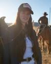 Young woman in a denim jacket and white "austin, texas" cap standing in a sunlit field, with horseback riders in the background.