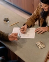 Two people sit at a counter; one holds a decorative belt buckle while the other sketches a design on paper. Several ornate buckles are displayed in an open box on the countertop.