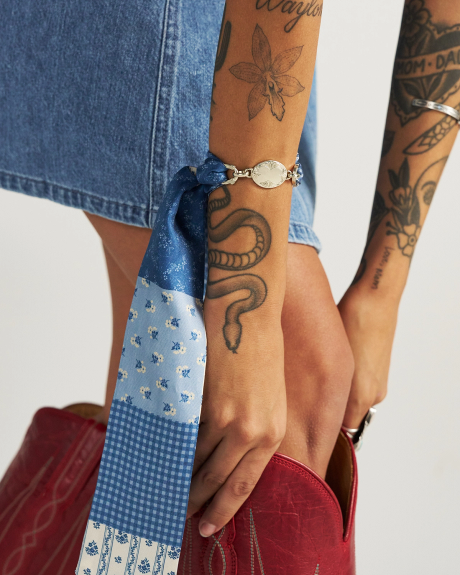 A person with tattooed arms holds a red cowboy boot, wearing a blue denim skirt and a blue patterned scarf tied at the wrist.