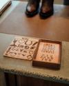 Two wooden trays on a counter: one with various engraved symbols and shapes, the other with engraved letters and numbers. A pair of brown shoes is partially visible in the background.