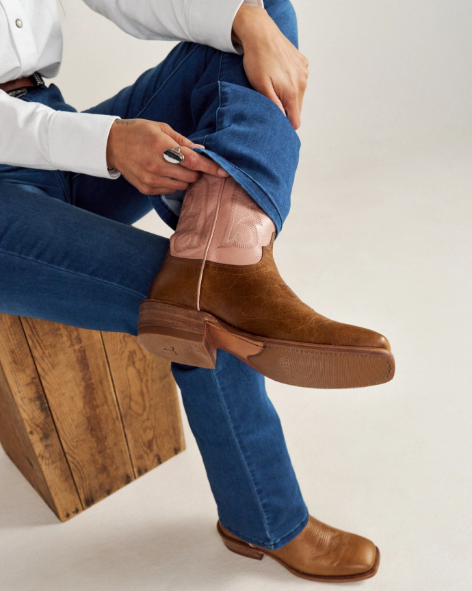 Person sitting on a wooden stool, wearing blue jeans and brown cowboy boots, adjusting the jeans over the boot.