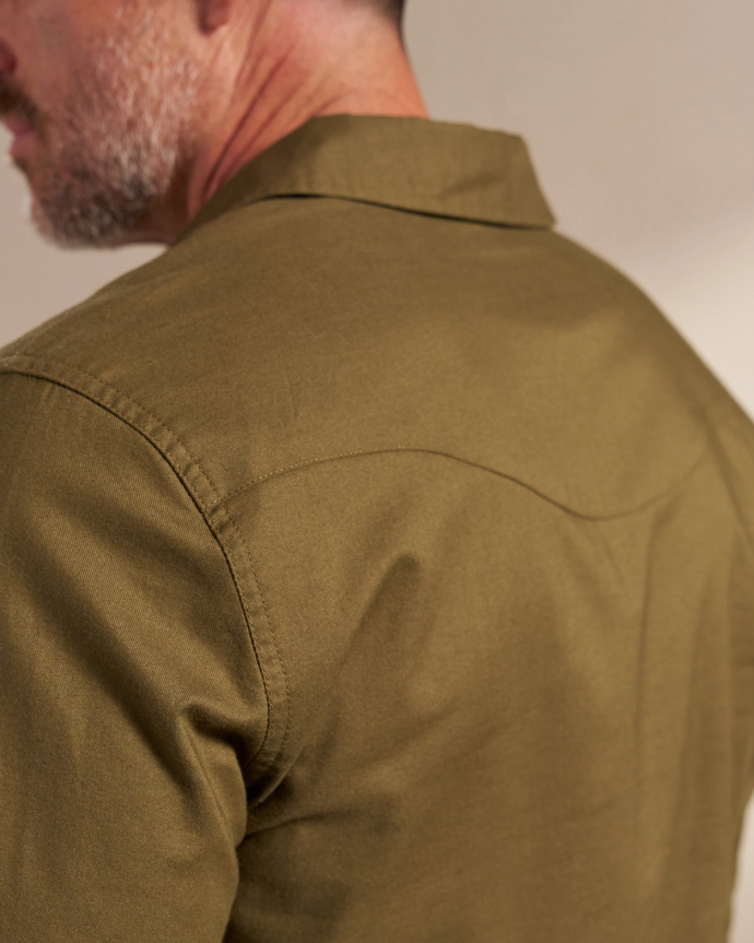 Close-up of a man in an olive green shirt, showing the back and shoulder area. The man has a short beard and mustache. The image is focused on the shirt's fabric and stitching details.