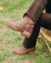 A person wearing brown boots and dark pants sits on a wicker chair on grass, with one leg crossed over the other.