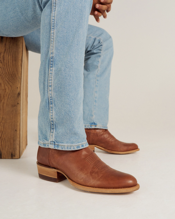 Person wearing light blue jeans and brown leather heeled boots sits on a wooden stool against a plain background.