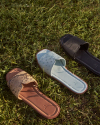 Three pairs of slip-on sandals lie on grass. The sandals are displayed with different designs: brown with a snake pattern, light blue with studs, and solid black.