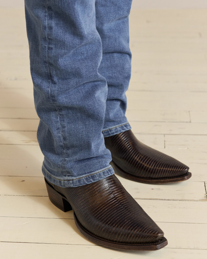 Person wearing blue jeans and dark brown cowboy boots stands on a light wooden floor.