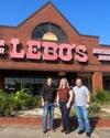 Three people stand in front of Lebo's store, which specializes in dance wear, footwear, and western wear. The building is brick with large signage.