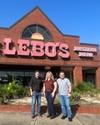 Three people stand in front of Lebo's store, which specializes in dance wear, footwear, and western wear. The building is brick with large signage.
