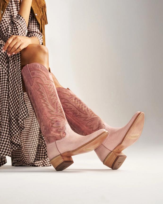 A person wearing a brown plaid dress and pink embroidered cowboy boots sits with legs crossed in front of a plain background.