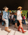 People at an outdoor event, wearing cowboy boots and hats, hold drinks. A woman in a white outfit and a woman in denim walk side by side. Background attendees are also in casual, western attire.