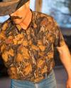 A person wearing a brown camouflage shirt, blue jeans, and a black cowboy hat with a large mustache, partially obscuring their face.