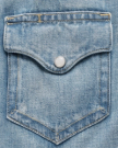 Close-up of a light blue denim jacket pocket with a silver snap button and visible yellow stitching.