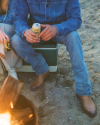 Two people sit on a cooler near a campfire, holding canned drinks. One wears cowboy boots and denim clothes, the other wears white boots and a striped skirt. Sand is visible on the ground.