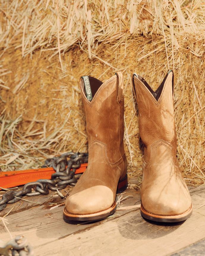A pair of tan cowboy boots placed on wooden planks in front of hay bales, with a chain and orange strap nearby.