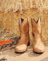A pair of tan cowboy boots placed on wooden planks in front of hay bales, with a chain and orange strap nearby.
