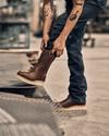 Person wearing brown boots and jeans, adjusting their pant leg in an industrial setting.