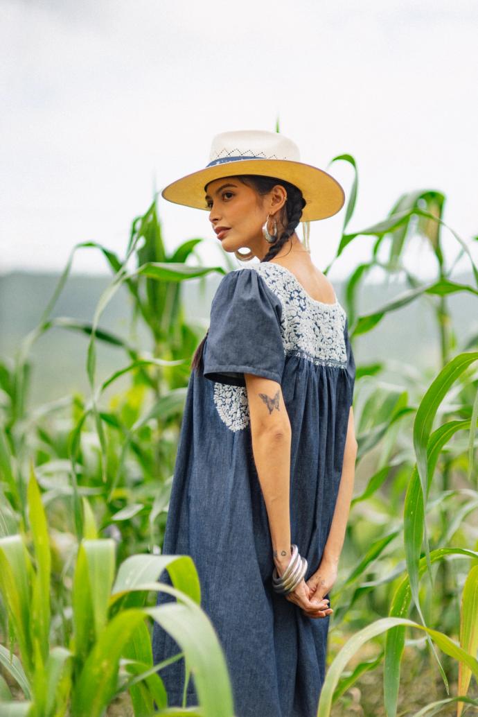 A woman in a wide-brimmed hat and blue dress stands in a field of tall plants, looking over her shoulder.