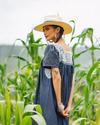 A woman in a wide-brimmed hat and blue dress stands in a field of tall plants, looking over her shoulder.