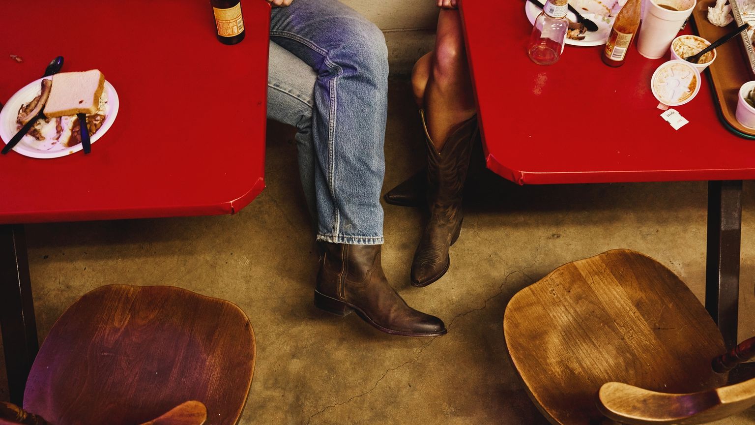 Two people wearing jeans and boots sit at red tables with leftover food and drinks; empty wooden chairs face them on a concrete floor.